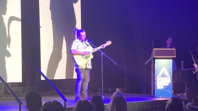 Man on stage playing guitar to large auditorium audience