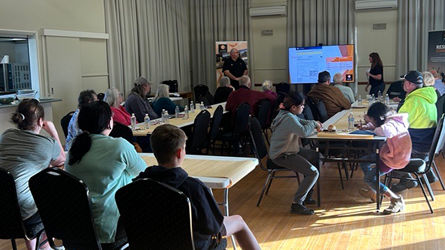 A group of residents sitting at trestle tables inside a small town hall watching a presentation