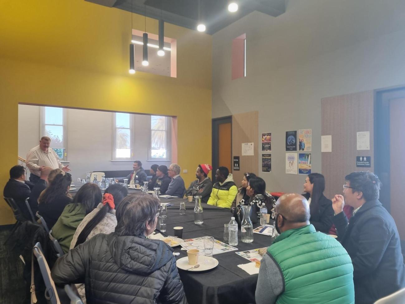 A group of 20 people sitting at a long lunch table, listening to a man giving a speech.