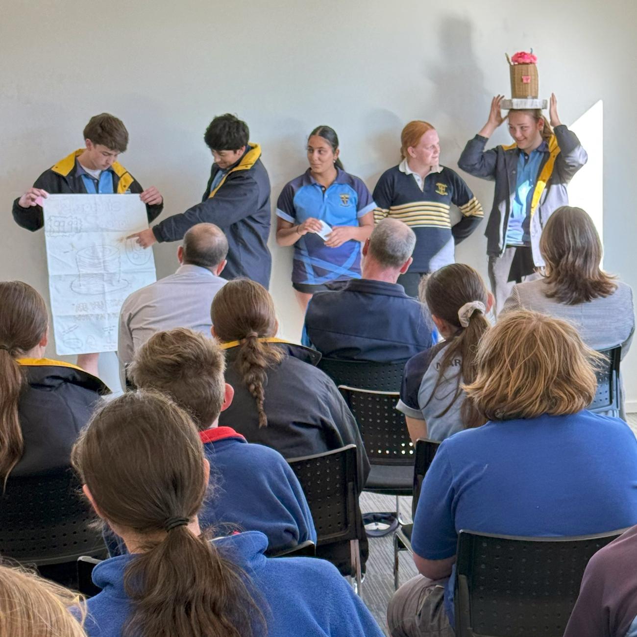 A group of high school students standing in front of an audience to give a presentation, one wearing a cardboard top hat