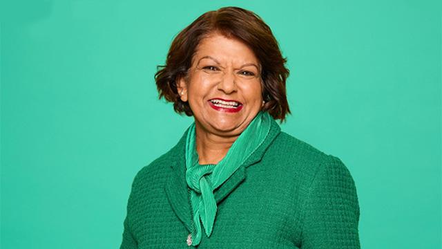 Senior lady in red lipstick, green scarf and green jacket smiling against a green background