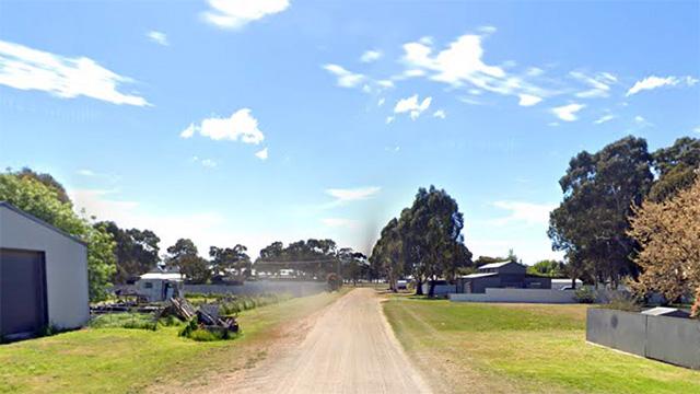 A rural dirt road with sunny sky and sparse buildings by roadside