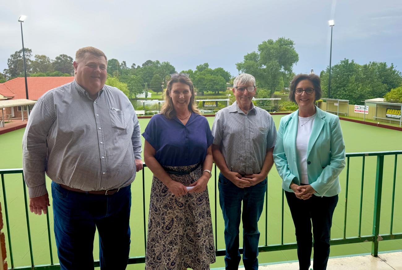 Four people standing on a balcony overlooking a lawn bowling green with a lake and fountain in the background