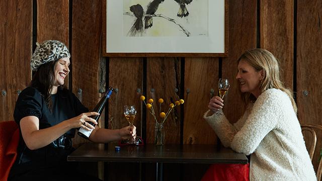 Two women sitting at a cafe table smiling and cheersing wine glasses