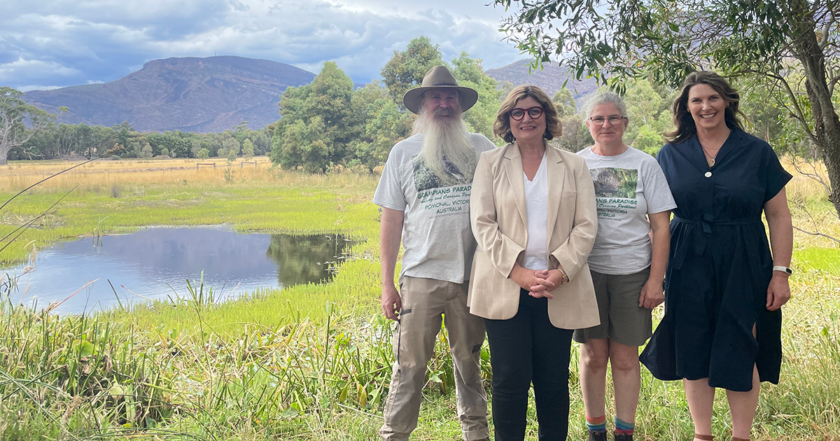 Four people stand together outdoors in front of a small pond, with trees and mountains in the background.