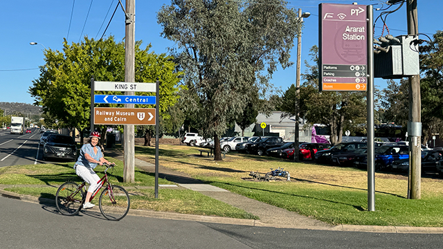 A bright, sunlit street with a woman wearing white pants riding a bicycle riding. She is smiling and looking into the camera